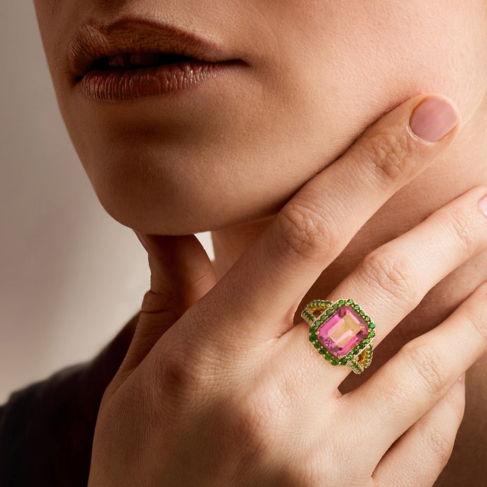 Ring on finger with pink square-cut gemstone and green halo stones in gold-tone