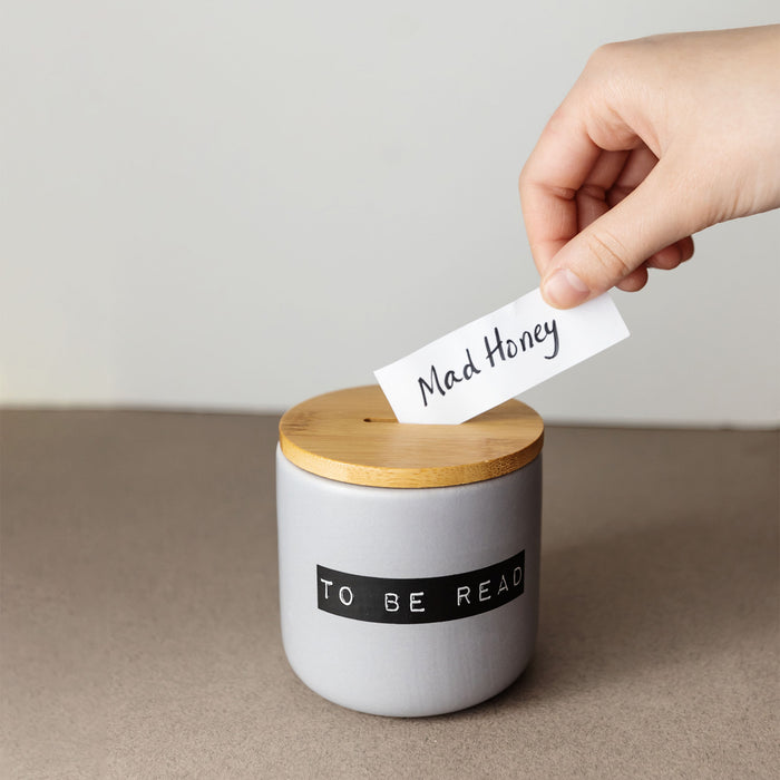 A hand places a "Mad Honey" slip into a ceramic "To Be Read" jar with a wooden lid.