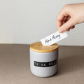 A hand places a "Mad Honey" slip into a ceramic "To Be Read" jar with a wooden lid.