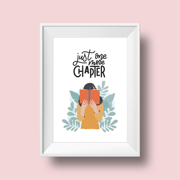 An art print hangs on a pink wall inside a white matted frame. The print features a white colored background with the words "Just one more chapter" written in black script over an illustration of a brunette girl holding an orange covered book in front of her face. Leaves stick out from behind her yellow shirt.
