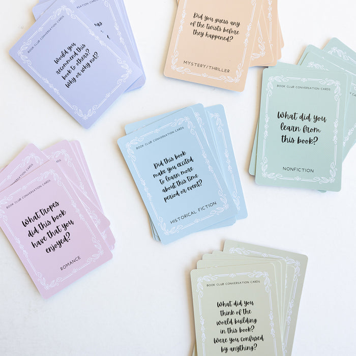 Five stacks of pastel Book Club Conversation Cards: purple cards, pink romance cards, blue historical fiction cards, orange mystery/thriller cards, green nonfiction cards, and olive cards. Each card has a different question on it.