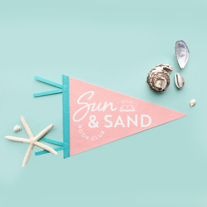 A light pink pendant flag that says Sun & Sand Book Club in white lettering with an icon of a book. The flag has two blue tassels sticking out the side and the flag is surrounded by shells.