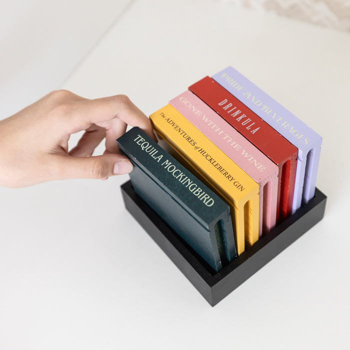 A hand pulls from a set of five drink coasters featuring drink puns from classic novels including "Drinkula", "Tequila Mockingbird", "Gone with the Wine", "The Adventures of Huckleberry Gin", and "Pride and Beverages".