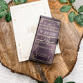 The front of a book-shaped phone case inspired by The Great Gatsby by F. Scott Fitzgerald. The phone case sits on a white book cover on a wood slice surrounded by greenery.