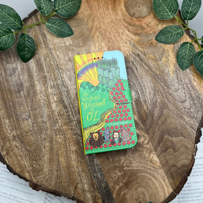 The front cover of a book-shaped phone case inspired by The Wonderful Wizard of Oz by L. Frank Baum. The phone case sits on a wood slice surrounded by greenery.
