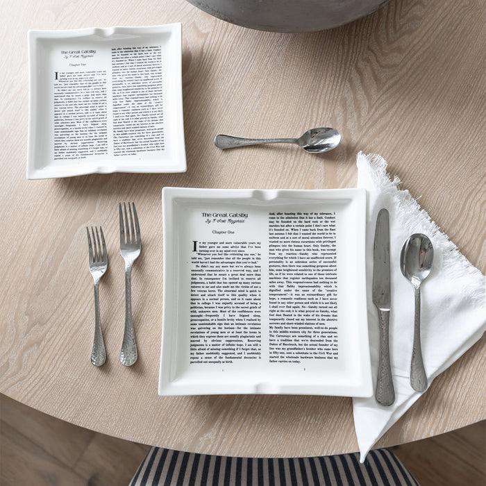 Two dinner plates with The Great Gatsby text, paired with silver cutlery on a wooden dining table.