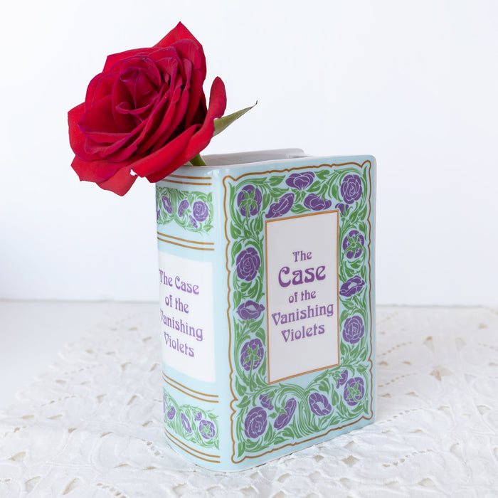 Book shaped vase labeled The Case of the Vanishing Violets in green and purple florals with a red rose sticking out of the top