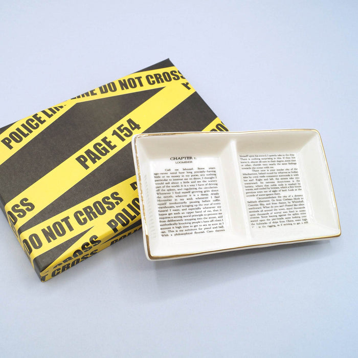 Ceramic ring dish printed with book text, placed beside a box wrapped in yellow "Police Line" tape.