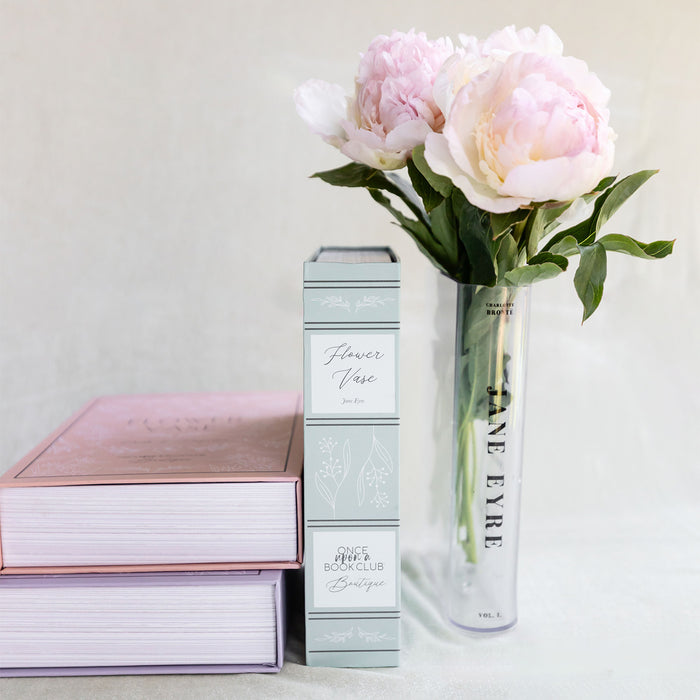 Pink and purple books stacked beside a tall glass vase labeled Jane Eyre, holding pale pink peonies.