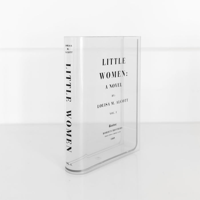 A clear acrylic book-shaped vase printed to look like a copy of Little Women by Louisa M. Alcott sits on a white surface. The spine of the 'book' faces outward and can be read in black bold font.
