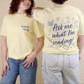 Two women wearing butter yellow t-shirts with 'Booked & Busy' on the front and 'Ask Me What I'm Reading' on the back surrounded by book-themed doodles.