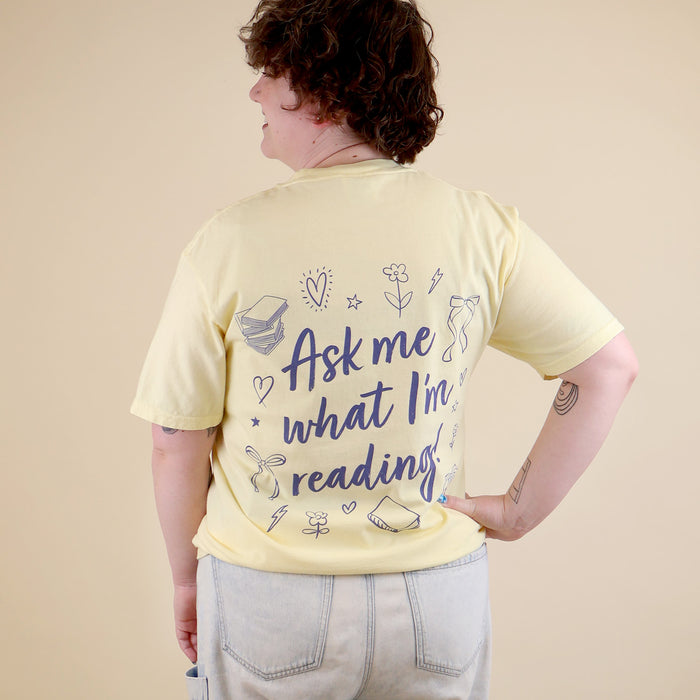 A woman wearing a butter yellow t-shirt with 'Booked & Busy' on the front and 'Ask Me What I'm Reading' on the back surrounded by book-themed doodles.