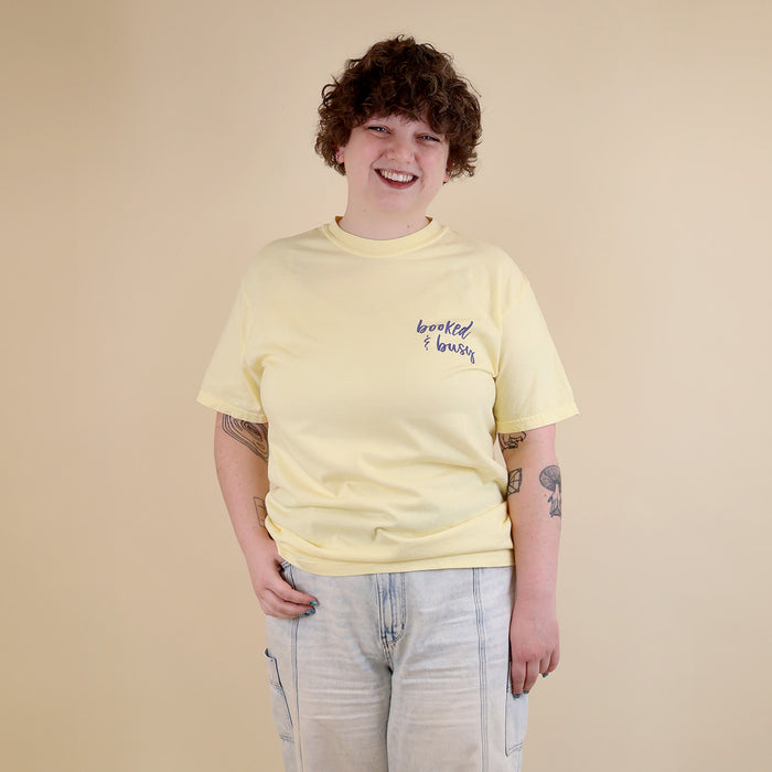 A woman wearing yellow t-shirts with 'Booked & Busy' on the front and 'Ask Me What I'm Reading' on the back surrounded by book-themed doodles.