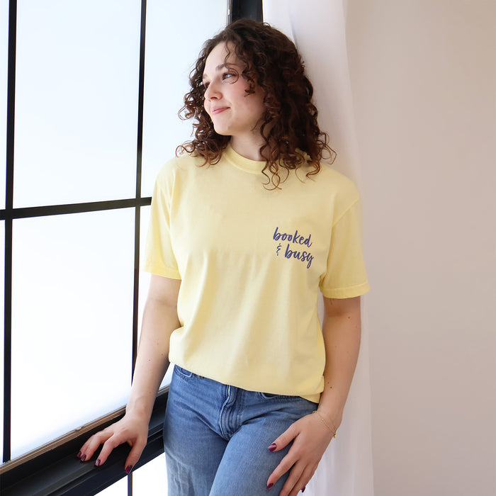A woman wearing yellow t-shirts with 'Booked & Busy' on the front and 'Ask Me What I'm Reading' on the back surrounded by book-themed doodles.