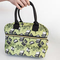 A hand holds a light green lunch box with black handles. There is a pattern of books and flowers across the green.