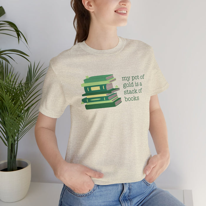 Person wearing a beige t-shirt with green stack of books and clovers graphic and text "my pot of gold is a stack of books"  standing next to a plant.