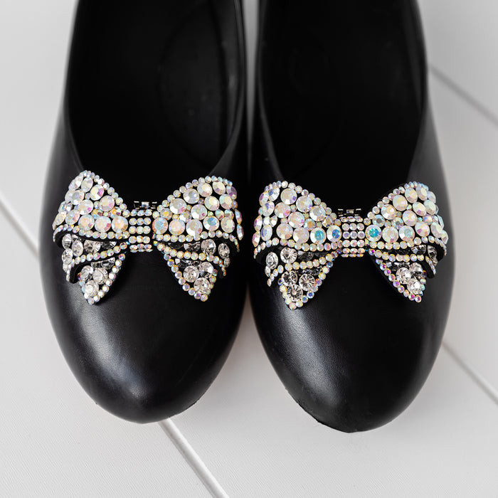 Black ballet flats have sparkling bow-shaped shoe clips attached to the fronts of them.