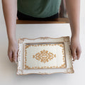 Hands holds out a white mirrored tray with gold accents over a white table.