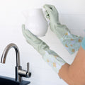 Hands wearing light green dish gloves hold a white ceramic pitcher over a stainless steel sink.