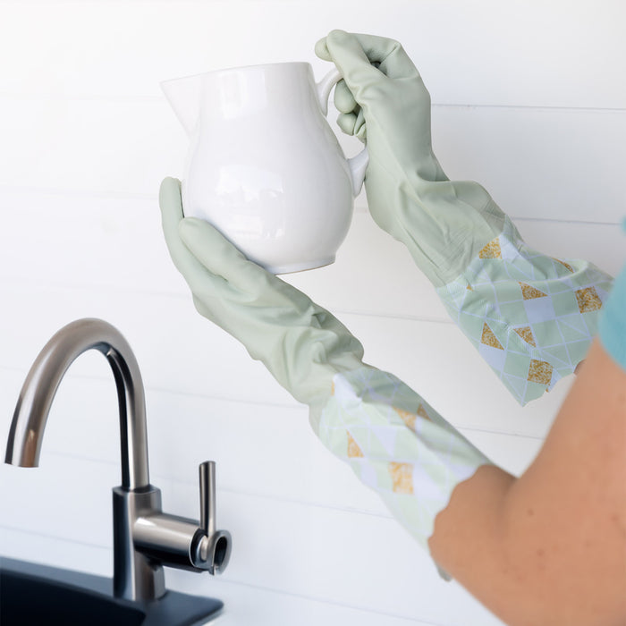 Hands wearing light green dish gloves hold a white ceramic pitcher over a stainless steel sink.