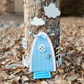 A light blue and white fairy door is attached to the trunk of a tree. White clouds are above it and a white picket fence is on each side. A small light blue welcome mat is in front.