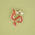 A gold dragon-shaped brooch with red sequins along the body, a green jewel eye, and a pearl in its mouth.