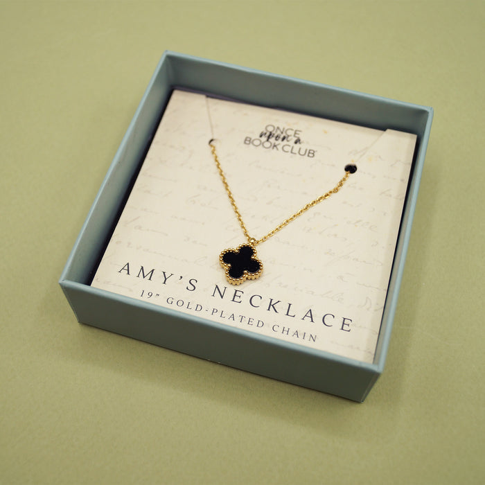 A small square open box with a gold-plated necklace. The necklace has a black cross-shaped charm on it.