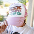 a woman wearing a pink snapback trucker hat that says "empowered women empower women" on the front, holding the brim of the hat