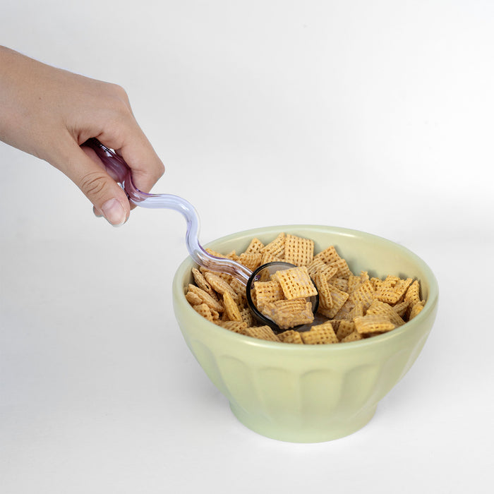 Hand using a purple spoon to scoop cereal from a green bowl on a white background