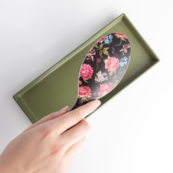 A black hairbrush with floral pattern in shades of pink, orange, blue, white, and green. The hairbrush is held over an open green box.