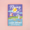 A hardcover copy of Flying Solo by Linda Holmes on a pink background.