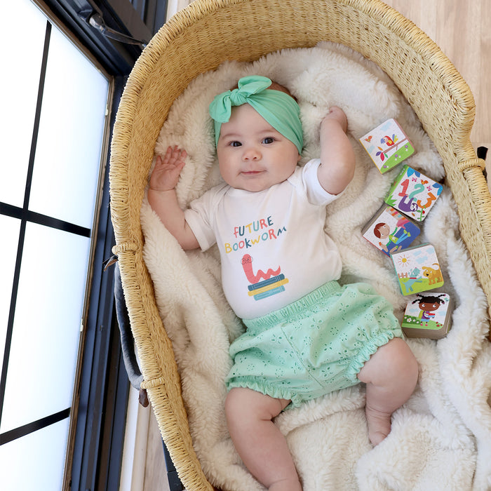 Baby in a bassinet wearing a 'Future Bookworm' shirt and green headband, surrounded by children's books.