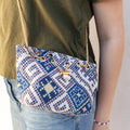 A round bubble purse with a woven, geometric pattern and gold chain strap is worn by a woman wearing jeans and a green shirt. Purse is closed with a gold chain slider clasp with faux-pearl details.
