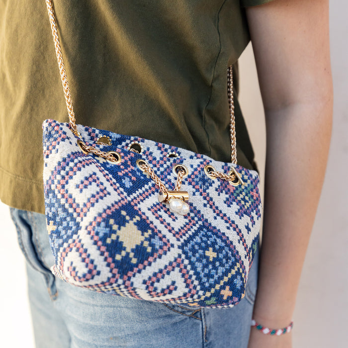A round bubble purse with a woven, geometric pattern and gold chain strap is worn by a woman wearing jeans and a green shirt. Purse is closed with a gold chain slider clasp with faux-pearl details.