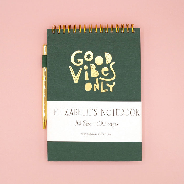 A dark green spiral notebook that says Good Vibes Only in gold lettering on the front. A gold pen is attached to the side.