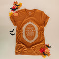 An orange-brown short-sleeve graphic T-shirt laid flat on a light beige background, surrounded by autumn leaves and small black bat cutouts. The shirt has a large cream decorative oval graphic on the front reading Harvest Moon Reader Society.