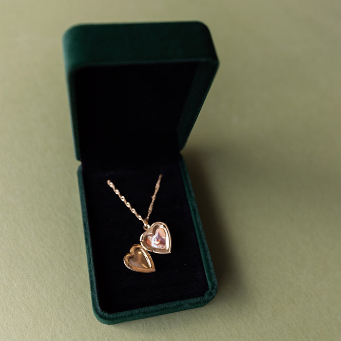 An open gold heart-shaped locked necklace with a photo of a cat inside, laying in a dark green jewelry case.