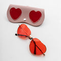 heart-shaped sunglasses with gold frames and red lenses next to a faux leather case with two red hearts on it