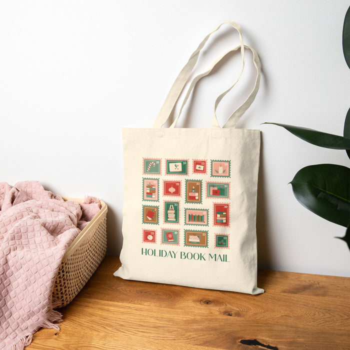 Tote bag with 'Holiday Book Mail' stamp design on a wooden surface with a plant in the background