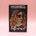 A paperback copy of Influence by Sara Shepard and Lilia Buckingham on a pink background.