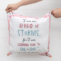 Two hands hold up a square pillow. The pillow is white with a light pink border with floral pattern on the edges. The quote in the middle says I am not afraid of storms, for I am learning how to sail my ship. Louisa May Alcott,  Little Women