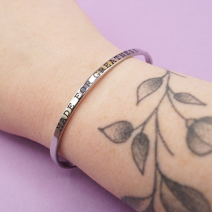 a silver cuff bracelet embossed with the words "Made for Greatness" is worn on someone's wrist.