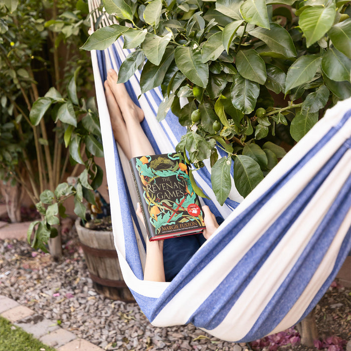 a blue and white striped hammock hung between two trees. Inside the hammock are a set of legs crossed at the ankle and a copy of The Revenant Games by Margie Fuston in their lap.