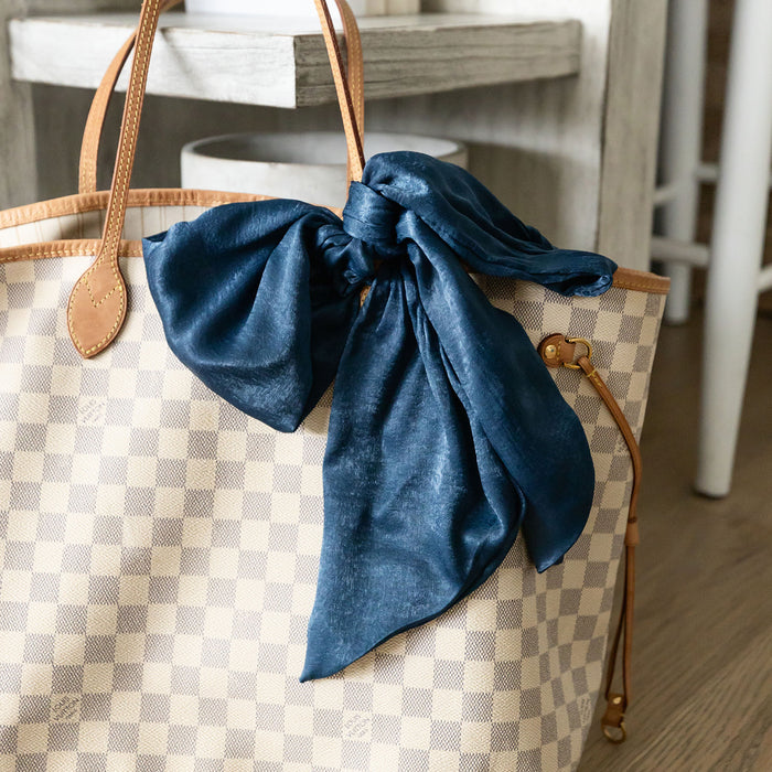 a dark blue skinny scarf tied in a bow around the handle of a checkered pattern handbag.