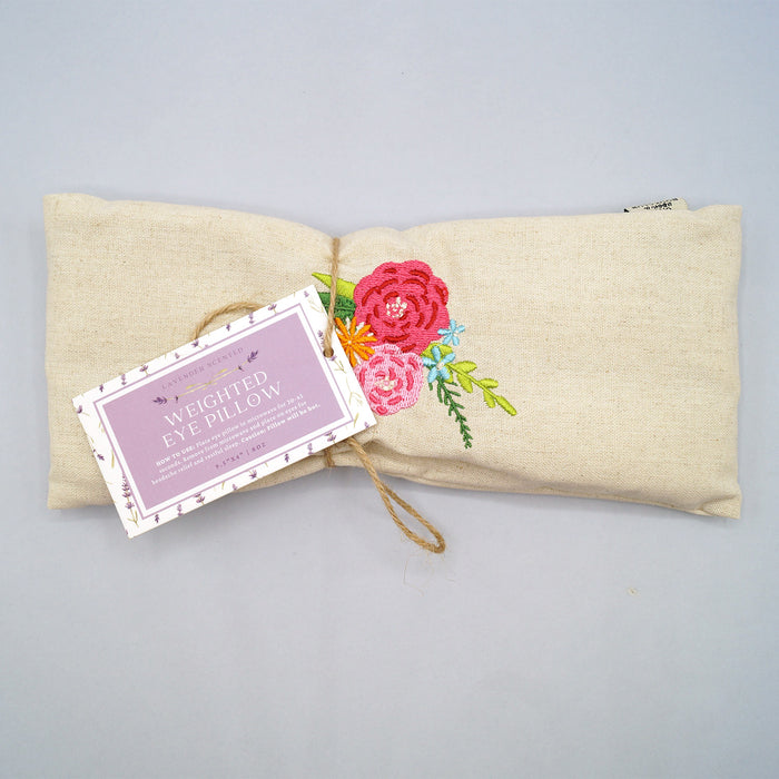 A beige weighted eye pillow with pink floral embroidery in the center.