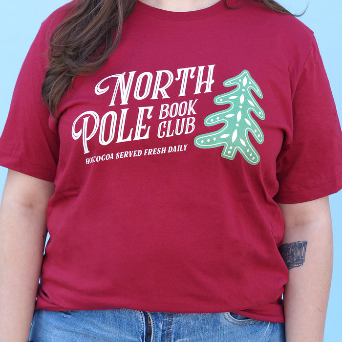 Red t-shirt with 'North Pole Book Club' text and green tree graphic on a blue background
