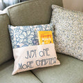 A square light tan pillow with light green/purple floral pattern lays on a couch. The front of the pillow has a pocket that covers the bottom half, with the words Just One More Chapter across it. A paperback edition of Hangry Hearts is tucked into the pocket.