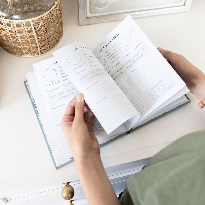 Two hands flip throughthe pages of the reading journal on a white countertop.