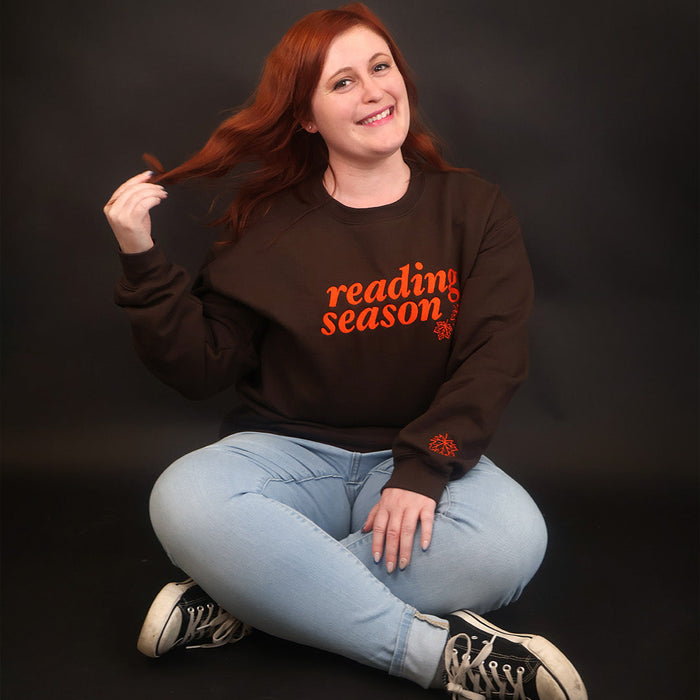 A smiling woman with red hair sits cross-legged on a black studio background, wearing a dark brown sweatshirt with orange text that reads 'reading season' and light blue jeans.