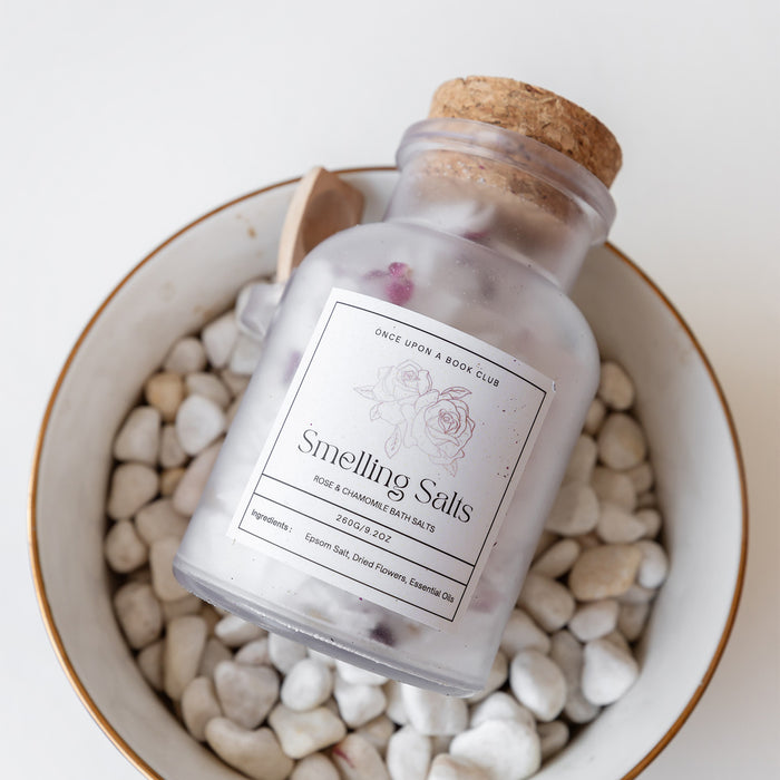 A jar with cork stopped labeled Smelling Salts - Rose and Chamomile Bath Salts - lays on a bowl filled with small white rocks.